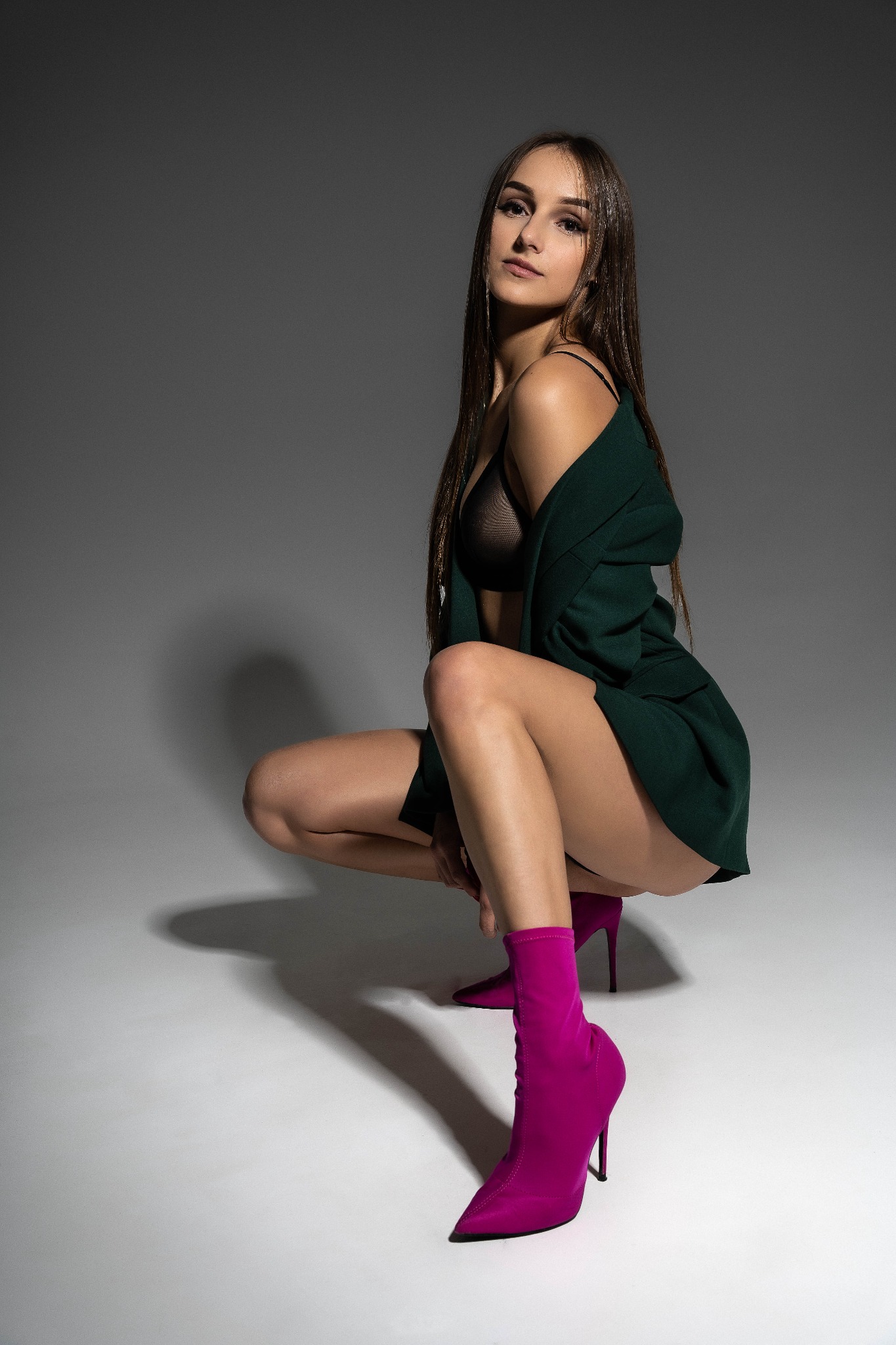 Kobieta w czarnej bieliźnie i zielonej marynarce, kucająca na białym tle, ubrana w różowe botki na obcasie, patrzy w obiektyw.