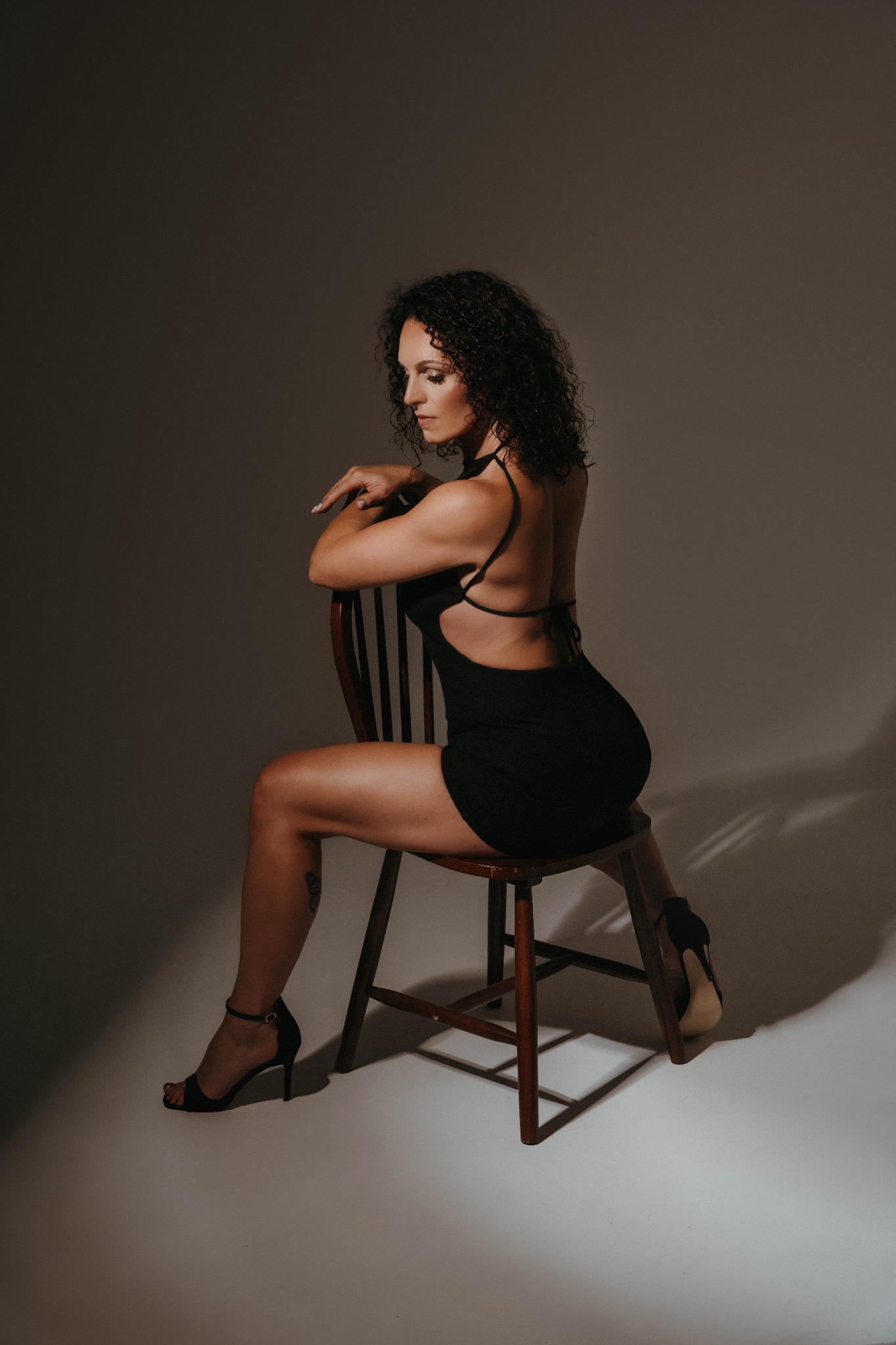 Kobieta z kręconymi włosami w czarnej sukience i szpilkach siedzi bokiem na drewnianym krześle, spoglądając w dół, w studiu z delikatnym oświetleniem.