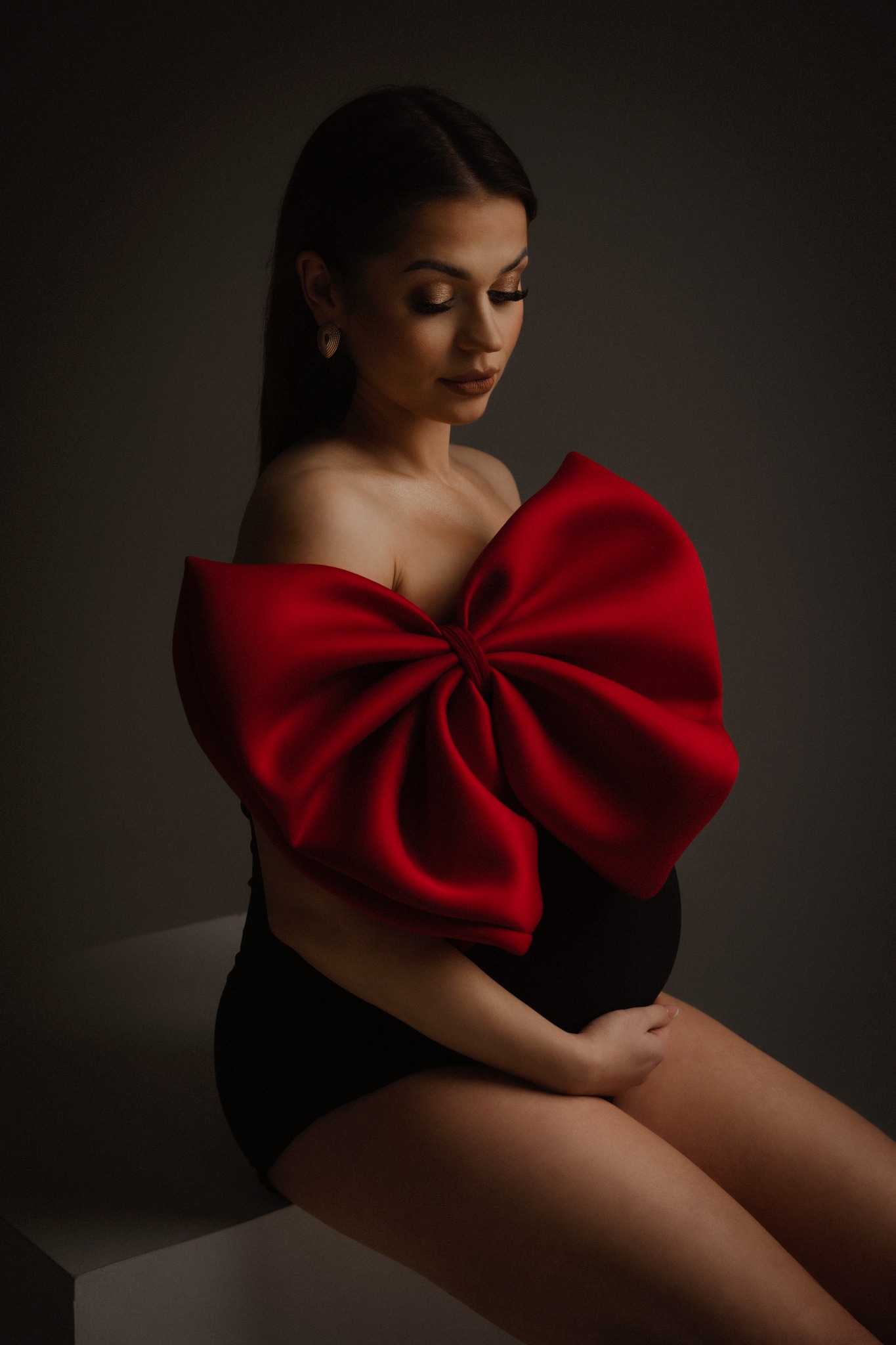 Portret kobiety w ciąży, ubrana w czarne body i ozdobiona dużą, czerwoną kokardą, delikatnie spoglądająca w dół i trzymająca dłoń na brzuchu w studiu z szarym tłem.