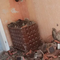 Fragment ceramicznego pieca kaflowego w trakcie rozbiórki, z widocznymi fragmentami kafli i gruzu na podłodze w pomieszczeniu z pomalowanymi ścianami i widocznymi śladami po instalacjach.