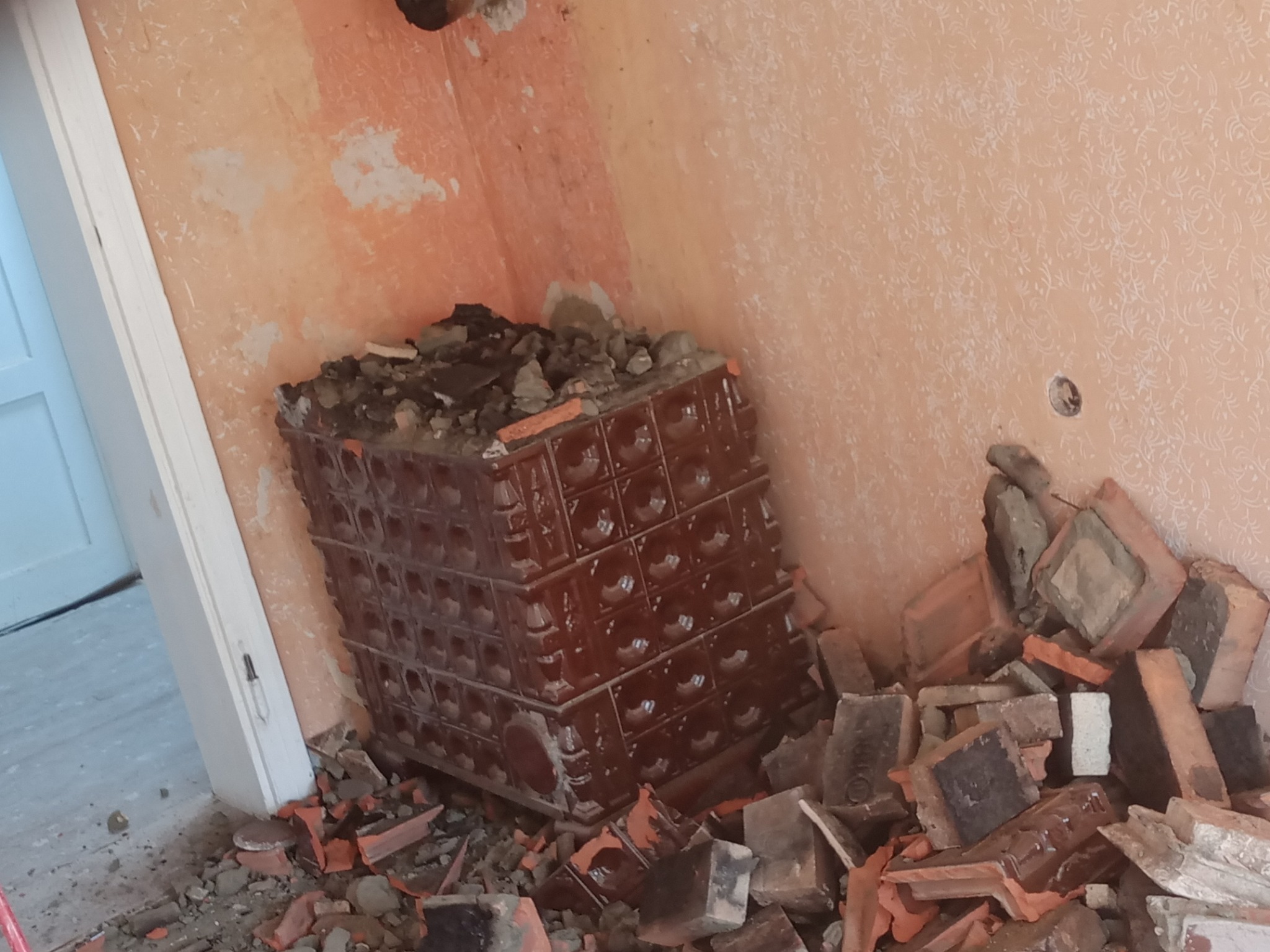 Fragment ceramicznego pieca kaflowego w trakcie rozbiórki, z widocznymi fragmentami kafli i gruzu na podłodze w pomieszczeniu z pomalowanymi ścianami i widocznymi śladami po instalacjach.