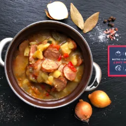 Zupa gulaszowa z kiełbasą i ziemniakami w metalowym garnku na czarnym tle, obok cebula, liście laurowe, pieprz i wizytówka firmy cateringowej.