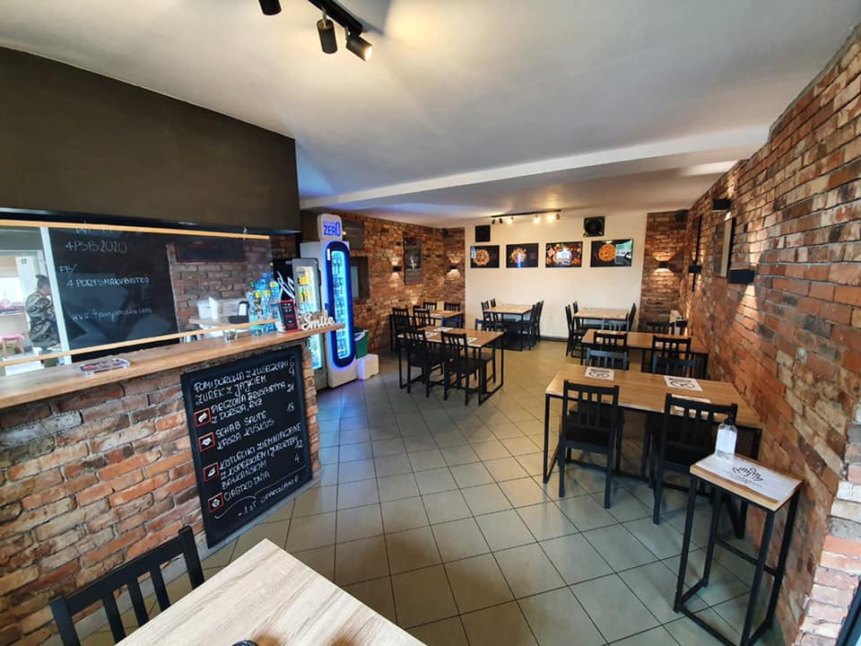 Wnętrze restauracji z ceglanymi ścianami, drewnianymi stołami i czarnymi krzesłami, menu wypisane kredą na tablicy, lodówka z napojami w tle.