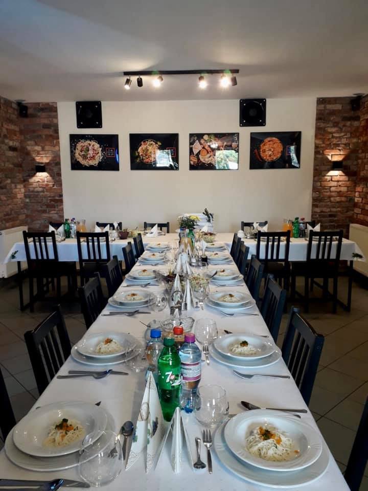 Długi stół nakryty białym obrusem z zastawą dla wielu gości, talerze z daniem na bazie makaronu, szklanki, sztućce, butelki z napojami, w tle ściana z cegły i zdjęcia potraw.