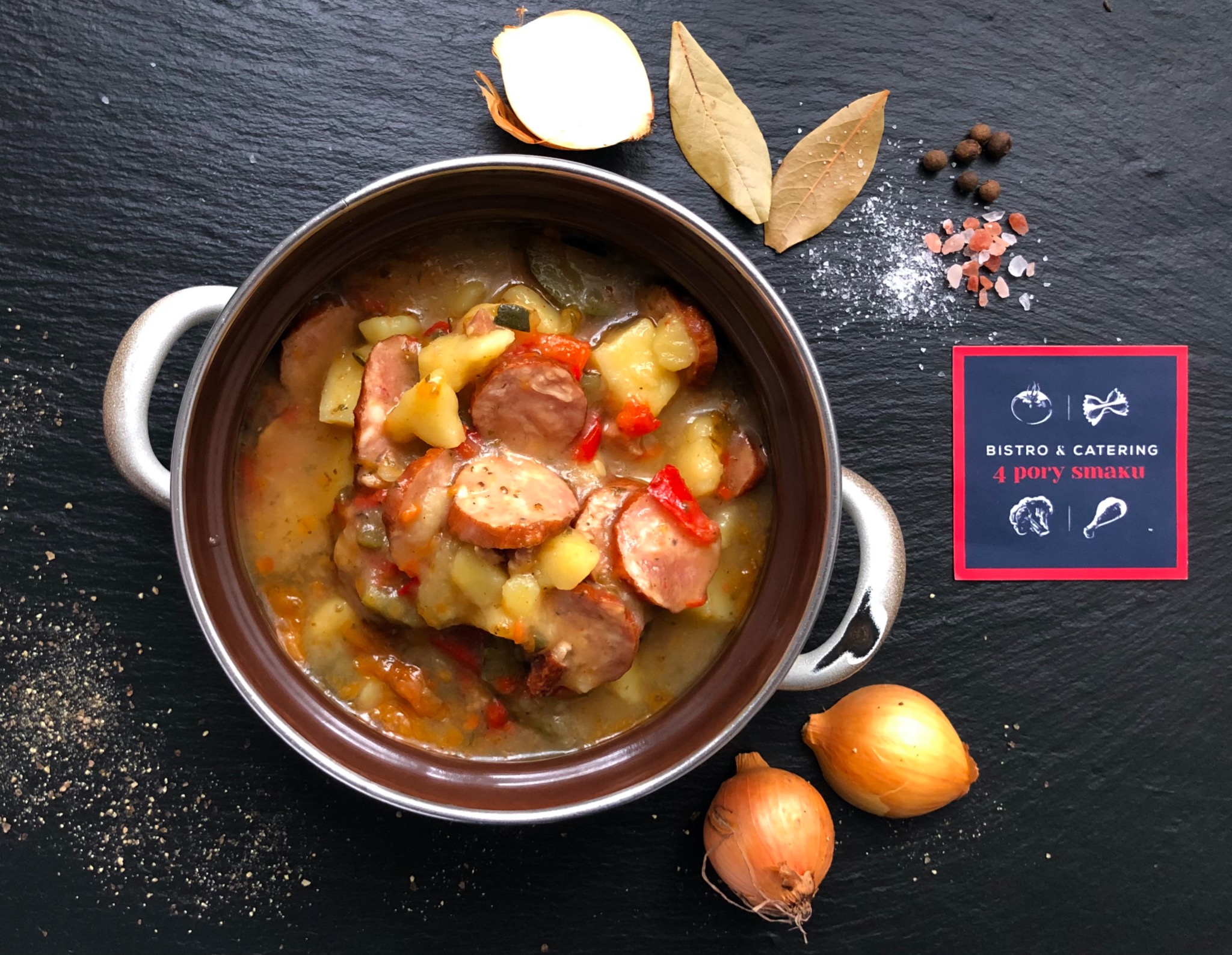 Zupa gulaszowa z kiełbasą i ziemniakami w metalowym garnku na czarnym tle, obok cebula, liście laurowe, pieprz i wizytówka firmy cateringowej.