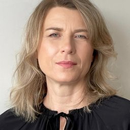 INON SPÓŁKA Z OGRANICZONĄ ODPOWIEDZIALNOŚCIĄ - Portret kobiety o jasnych włosach, ubrana w czarną bluzkę z wiązaniem, na neutralnym tle. Profesjonalny wizerunek.