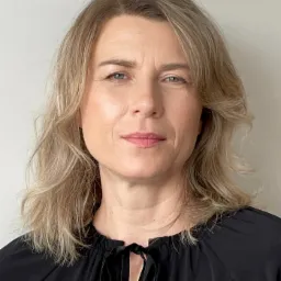 Portret kobiety o jasnych włosach, ubrana w czarną bluzkę z wiązaniem, na neutralnym tle. Profesjonalny wizerunek.