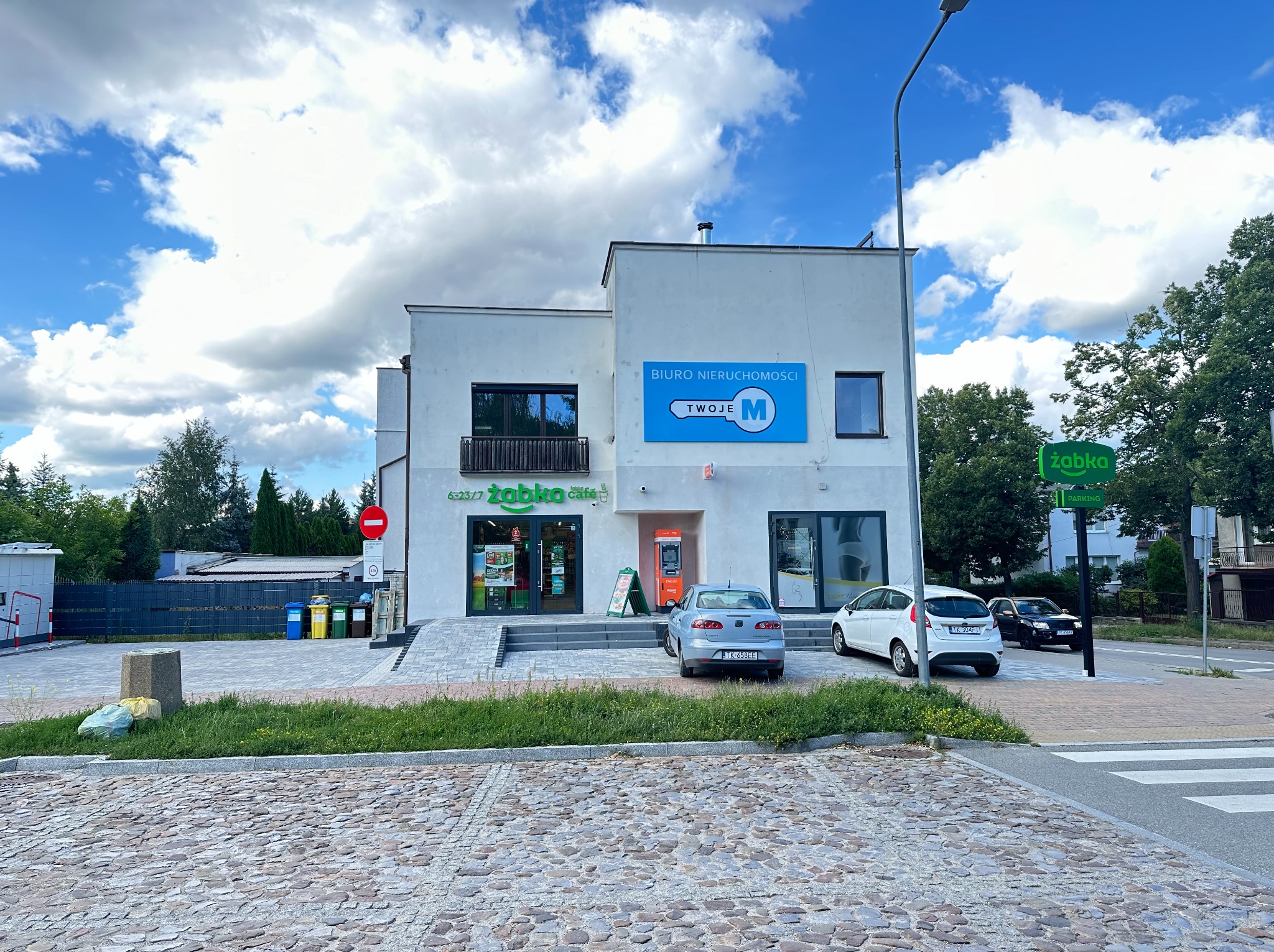 Biały budynek biura nieruchomości 'Twoje M' z szyldem, obok sklep Żabka, bankomat i zaparkowane auta. Ujęcie z poziomu ulicy, słoneczny dzień.