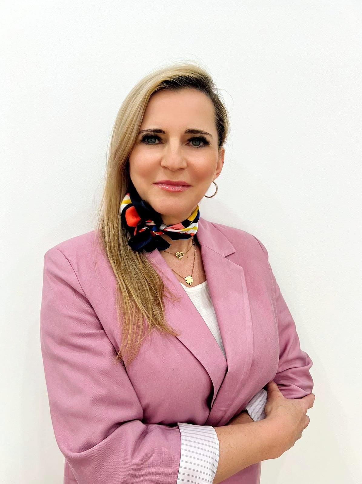 Profesjonalny portret kobiety w różowym żakiecie i wzorzystej apaszce, z uśmiechem i złotą biżuterią na tle białej ściany. Ujęcie biznesowe, budzące zaufanie.