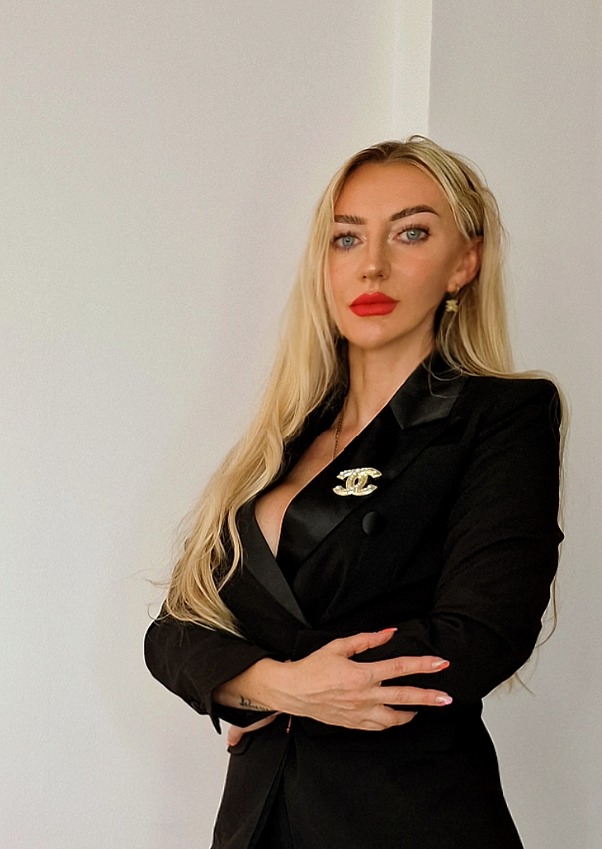 Profesjonalny wizerunek agentki nieruchomości: blondynka w czarnym garniturze z broszką Chanel, czerwone usta, na jasnym tle. Elegancki strój.