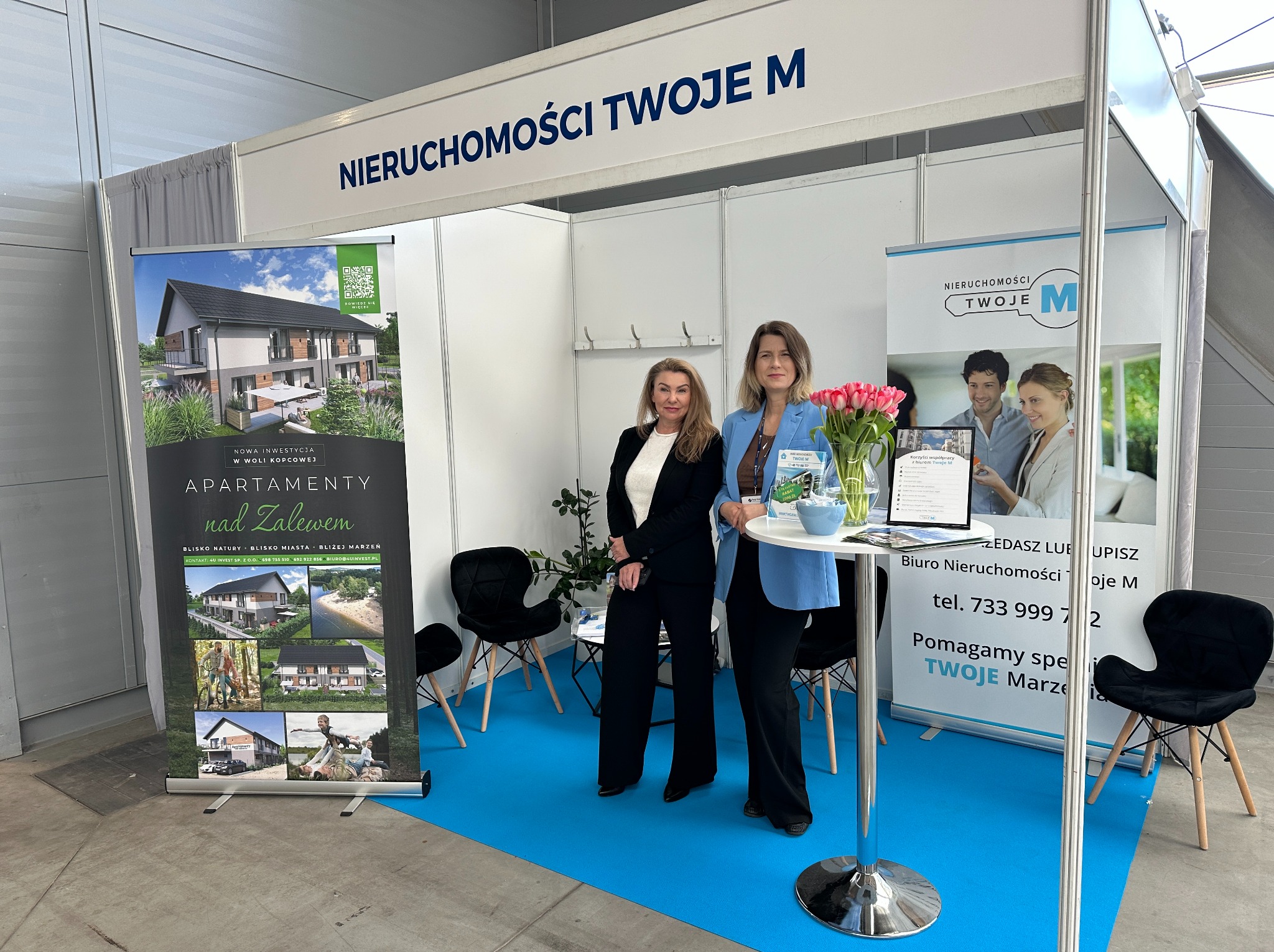 Stoisko targowe 'Nieruchomości Twoje M' z dwoma kobietami, prezentujące apartamenty nad Zalewem w Woli Kopcowej. Ekspozycja z wizualizacjami i danymi kontaktowymi.