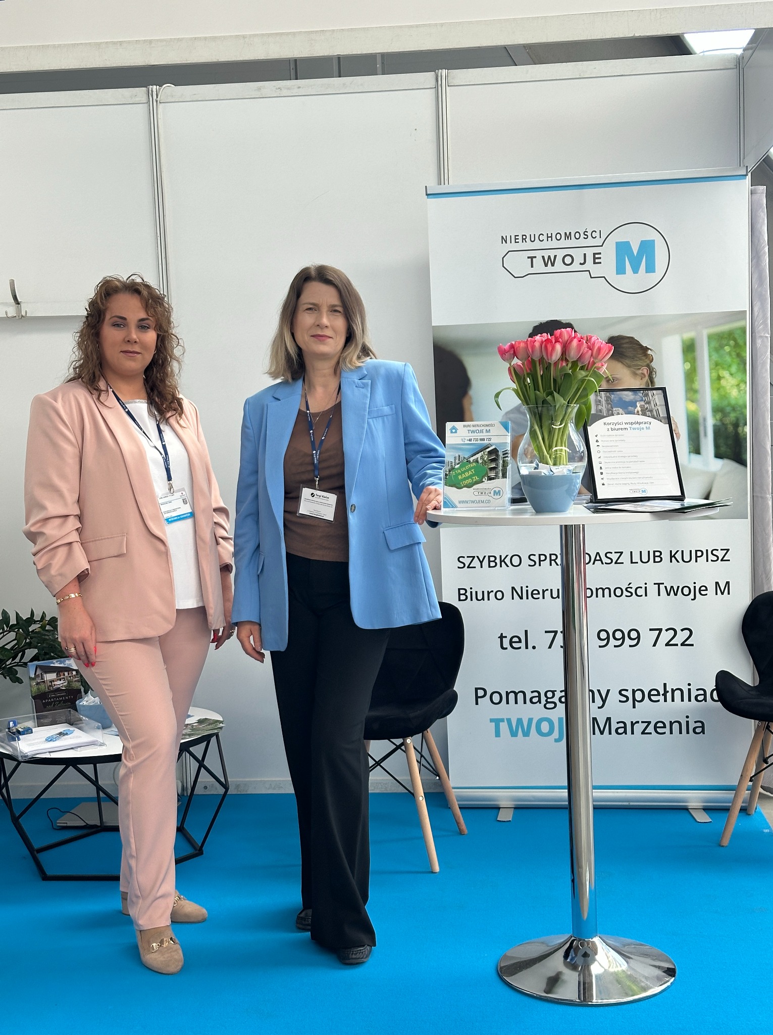 Dwie kobiety w eleganckich marynarkach prezentują ofertę biura nieruchomości 'Twoje M' na tle stoiska targowego z tulipanami i informacjami o ofercie.
