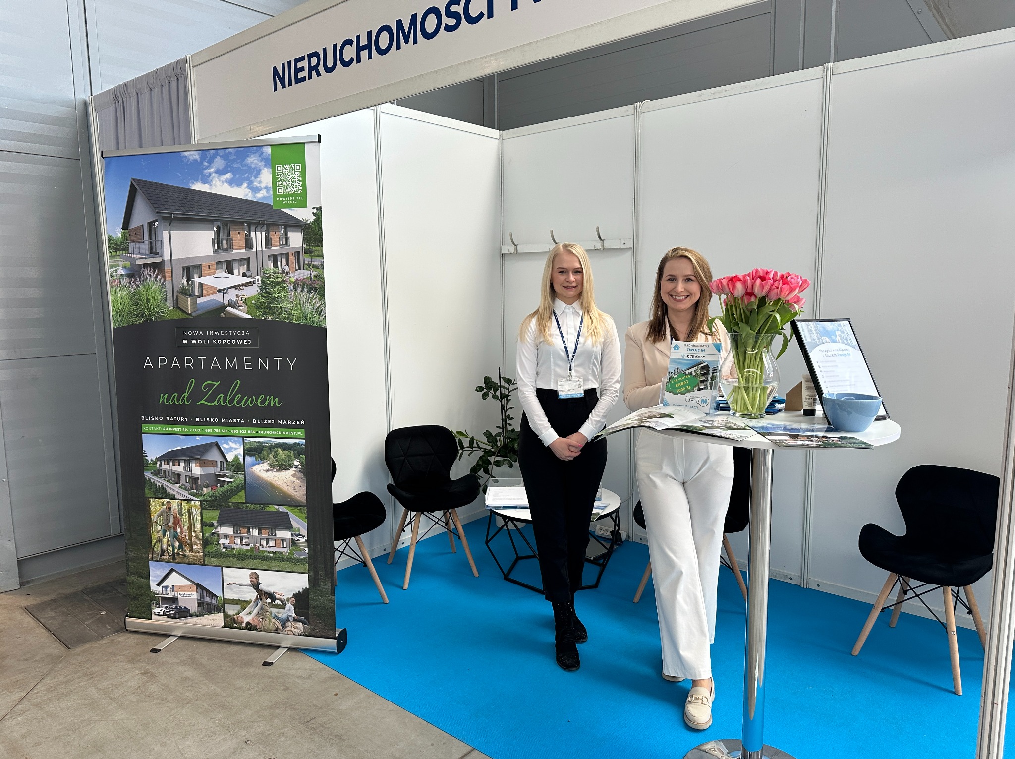 Stoisko targowe nieruchomości: dwie kobiety prezentują ofertę apartamentów nad Zalewem. Widoczny roll-up z wizualizacjami budynków i logo firmy.