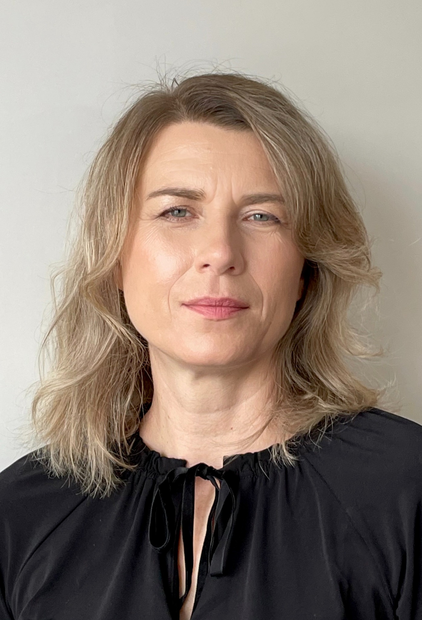 Portret kobiety o jasnych włosach, ubrana w czarną bluzkę z wiązaniem, na neutralnym tle. Profesjonalny wizerunek.