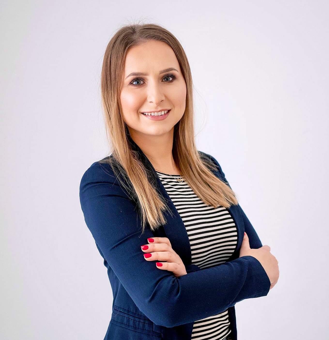 Uśmiechnięta blondynka w granatowej marynarce na jasnym tle, z rękami skrzyżowanymi na piersi. Profesjonalny wizerunek agenta nieruchomości.