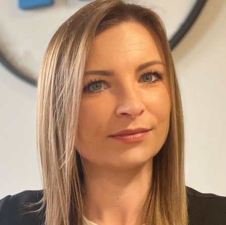 Portret kobiety z blond włosami, niebieskimi oczami i subtelnym uśmiechem, na rozmytym tle biurowym. Ujęcie z bliska, skupiające się na twarzy.