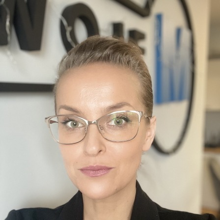 Portret kobiety w okularach na tle biura nieruchomości z logo. Profesjonalny wizerunek agenta nieruchomości, budzący zaufanie i kompetencje.