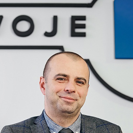 Uśmiechnięty mężczyzna w szarej marynarce na tle białej ściany z logo firmy w czarno-niebieskich barwach. Profesjonalny agent nieruchomości.