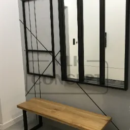 Nowoczesny przedpokój w Rajbrocie z drewnianą ławką na czarnych nogach, w tle lustro w czarnej ramie i wieszak na ubrania, całość dopełnia minimalistyczny wzór na ścianie.