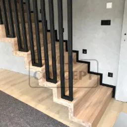 Nowoczesne schody z jasnego drewna z czarną, minimalistyczną balustradą z pionowych profili, oświetlenie punktowe w ścianie nad schodami.