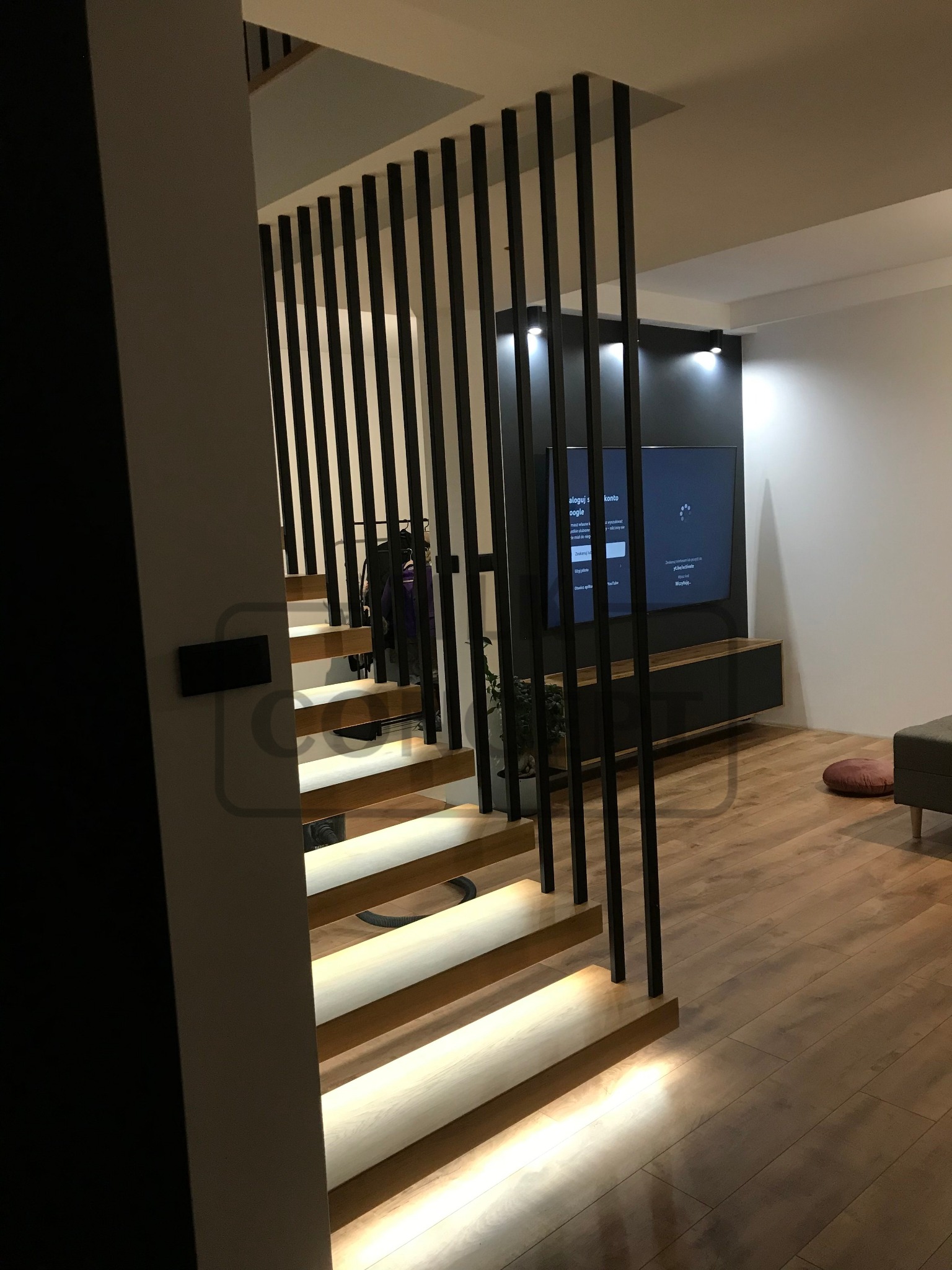 Nowoczesne schody z podświetlanymi stopniami i minimalistyczną balustradą z pionowych, czarnych prętów, na tle salonu z telewizorem.