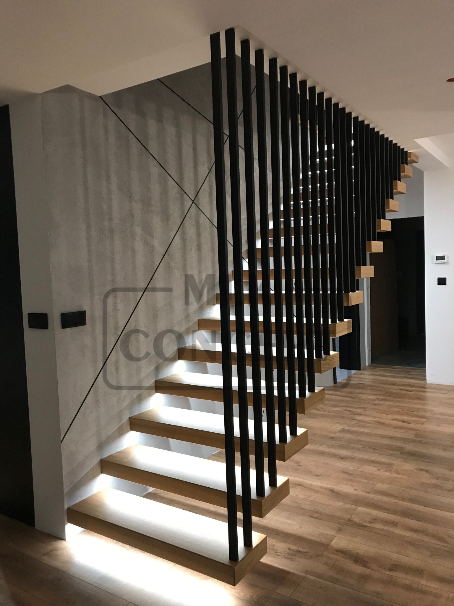 Nowoczesne schody z podświetlanymi stopniami i czarną, pionową balustradą, na tle ściany z geometrycznym wzorem i drewnianej podłogi.