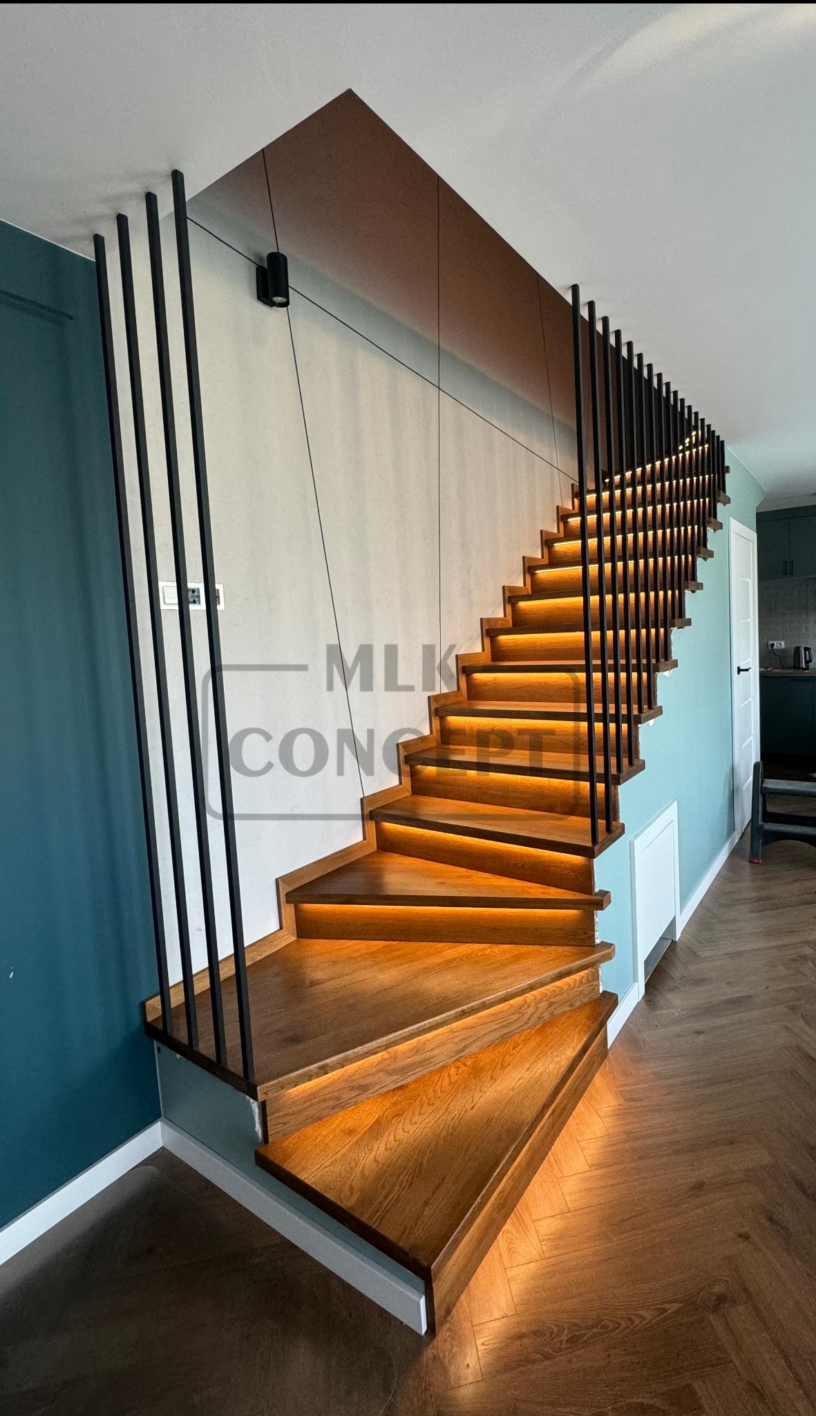 Drewniane schody z podświetleniem LED na każdym stopniu, balustrada z pionowych, czarnych prętów, ściana w kolorze morskim, podłoga ułożona w jodełkę.