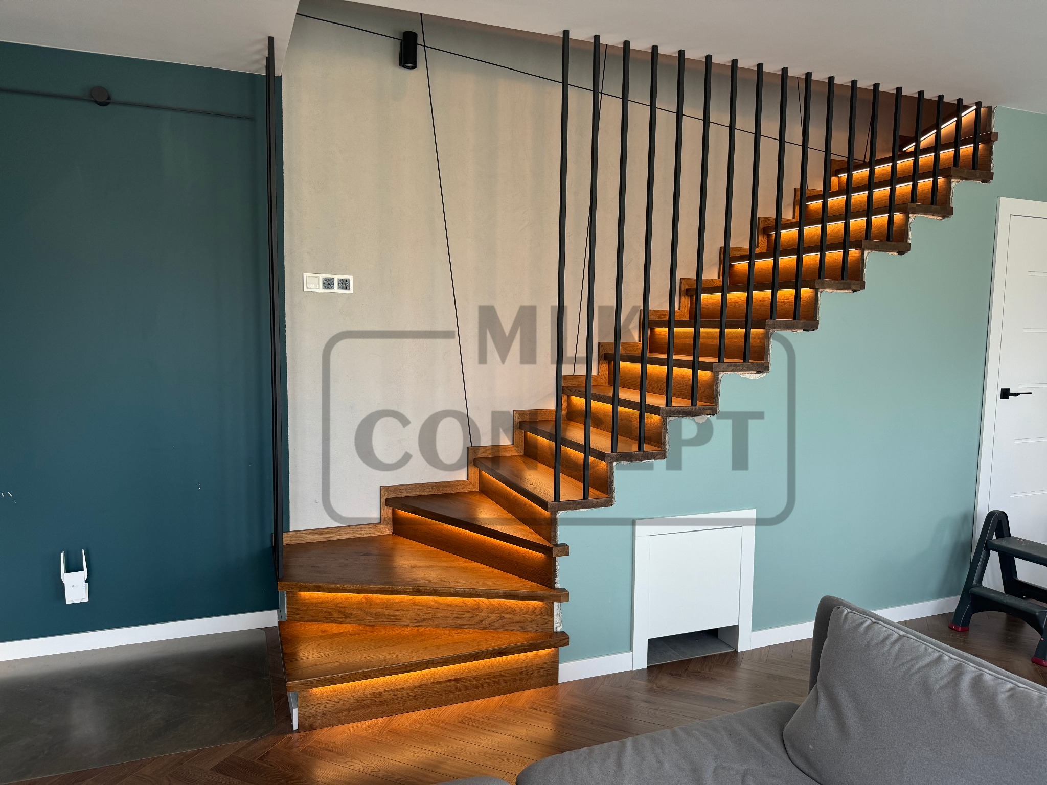 Nowoczesne schody z drewna z podświetleniem LED i czarnymi, pionowymi balustradami, widok z dołu, fragment szarej sofy w prawym dolnym rogu.