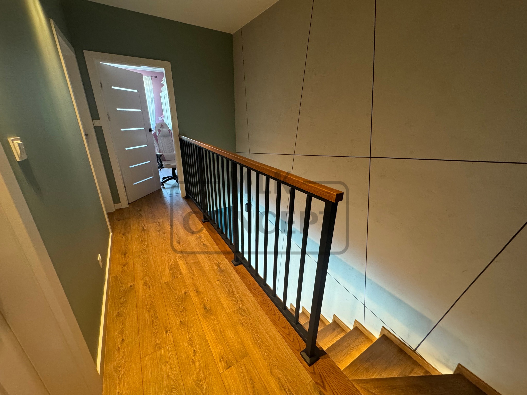 Schody drewniane z czarną balustradą i drewnianą poręczą, widok z góry na dół, z widocznym fragmentem podłogi i ściany w kolorze oliwkowym, oraz białymi drzwiami z poziomymi frezami.