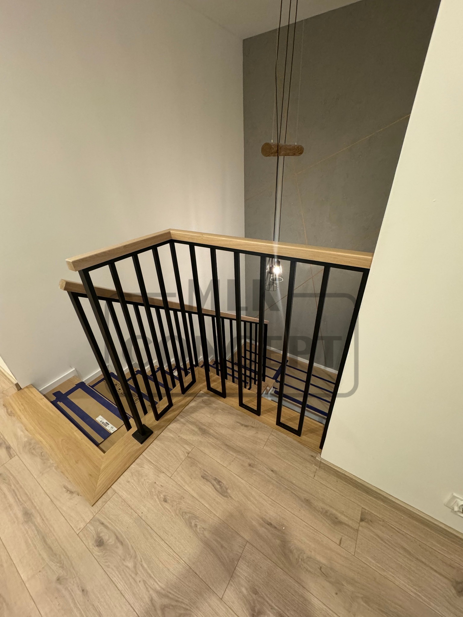 Kątowe ujęcie schodów z drewnianymi stopniami zabezpieczonymi kartonem i czarną balustradą z drewnianym poręczą, widok z góry na dół.