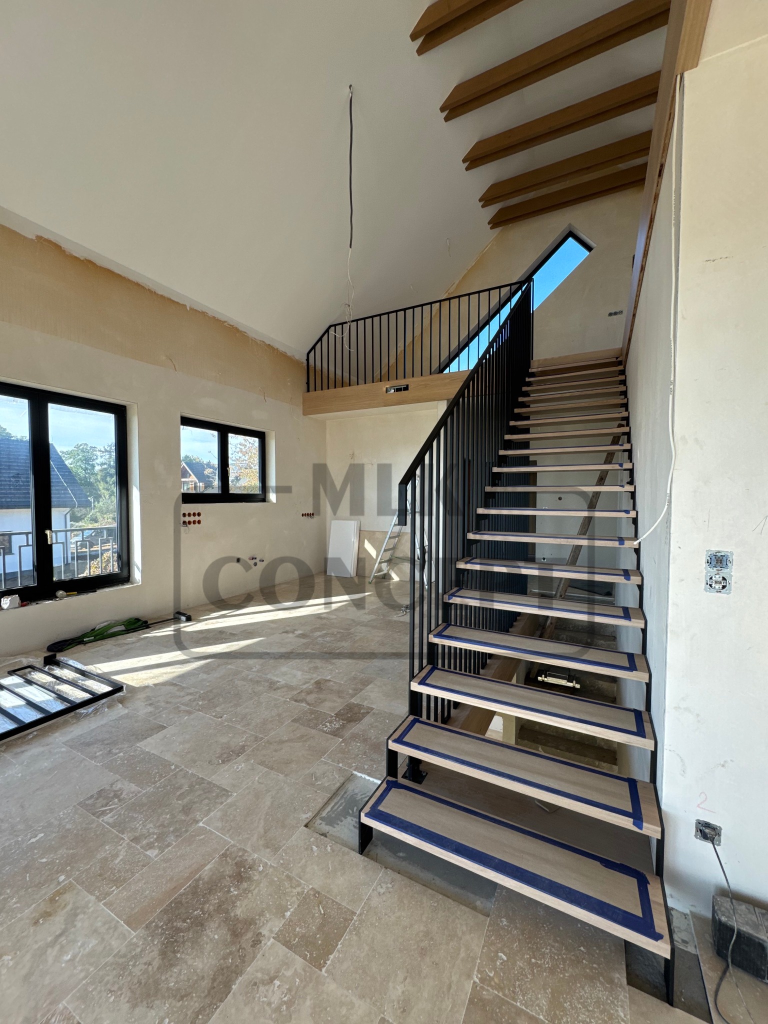 Wnętrze w trakcie wykończenia z drewnianymi schodami i czarną metalową balustradą. Schody zabezpieczone taśmą malarską, widoczne instalacje elektryczne i okna z widokiem na zewnątrz.