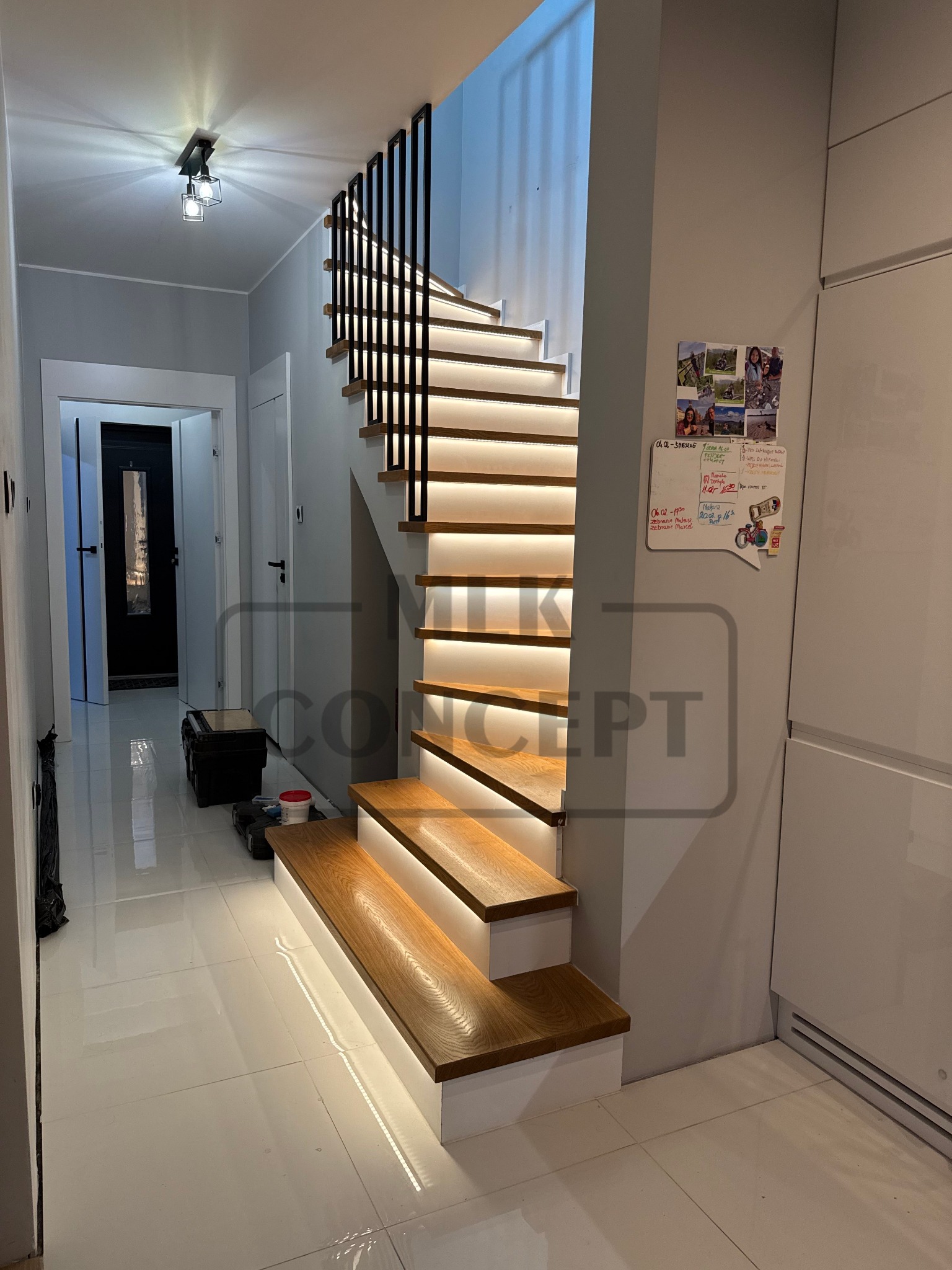 Nowoczesne schody z drewnianymi stopniami i podświetleniem LED, czarna balustrada z pionowych prętów, białe ściany i podłoga, widoczne drzwi w tle.