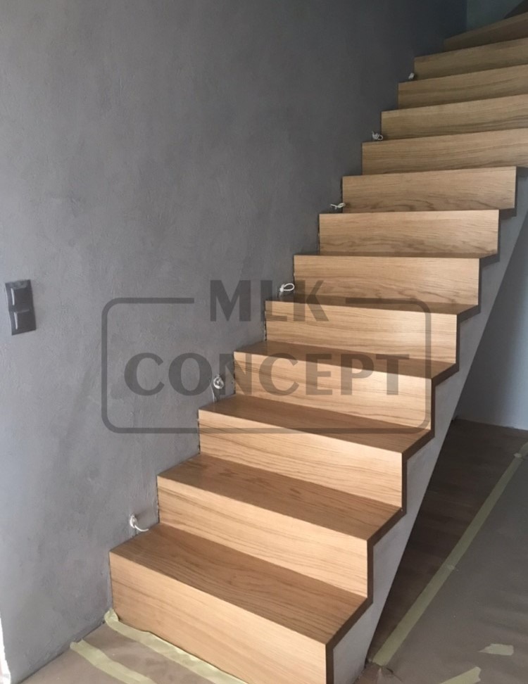 Drewniane schody o jasnym odcieniu drewna, zamontowane przy szarej ścianie, zabezpieczone taśmą malarską na podłodze, z widocznymi uchwytami montażowymi.