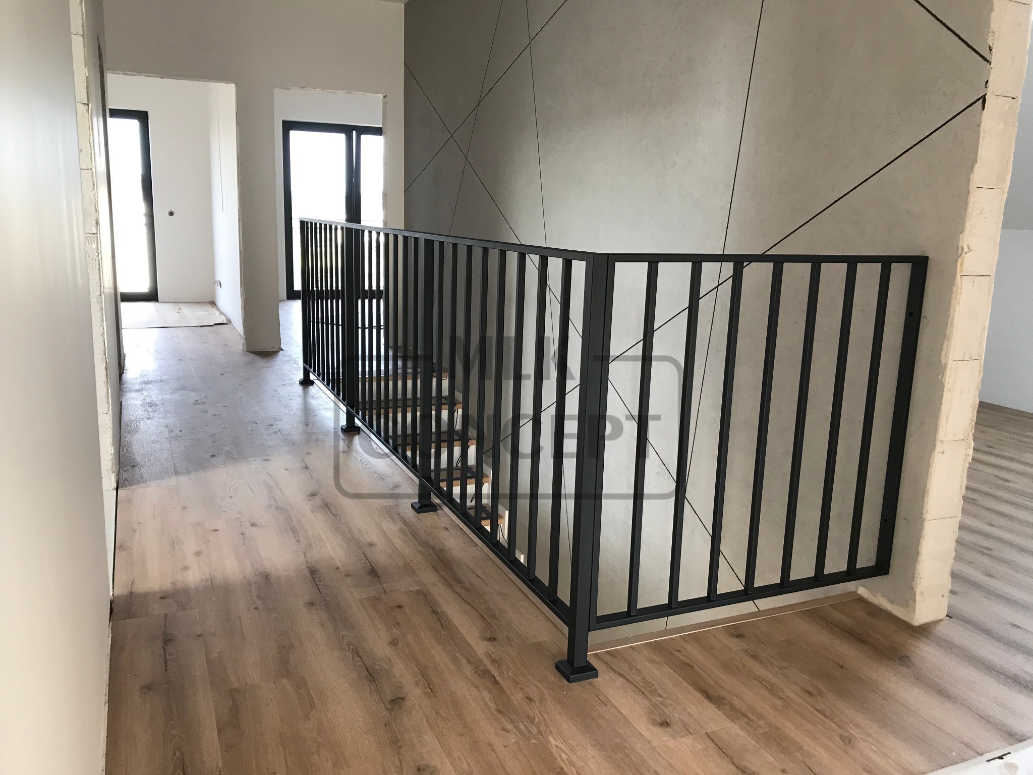 Nowoczesna czarna balustrada z pionowych prętów na schodach w jasnym wnętrzu z drewnianą podłogą i betonową ścianą z geometrycznym wzorem.