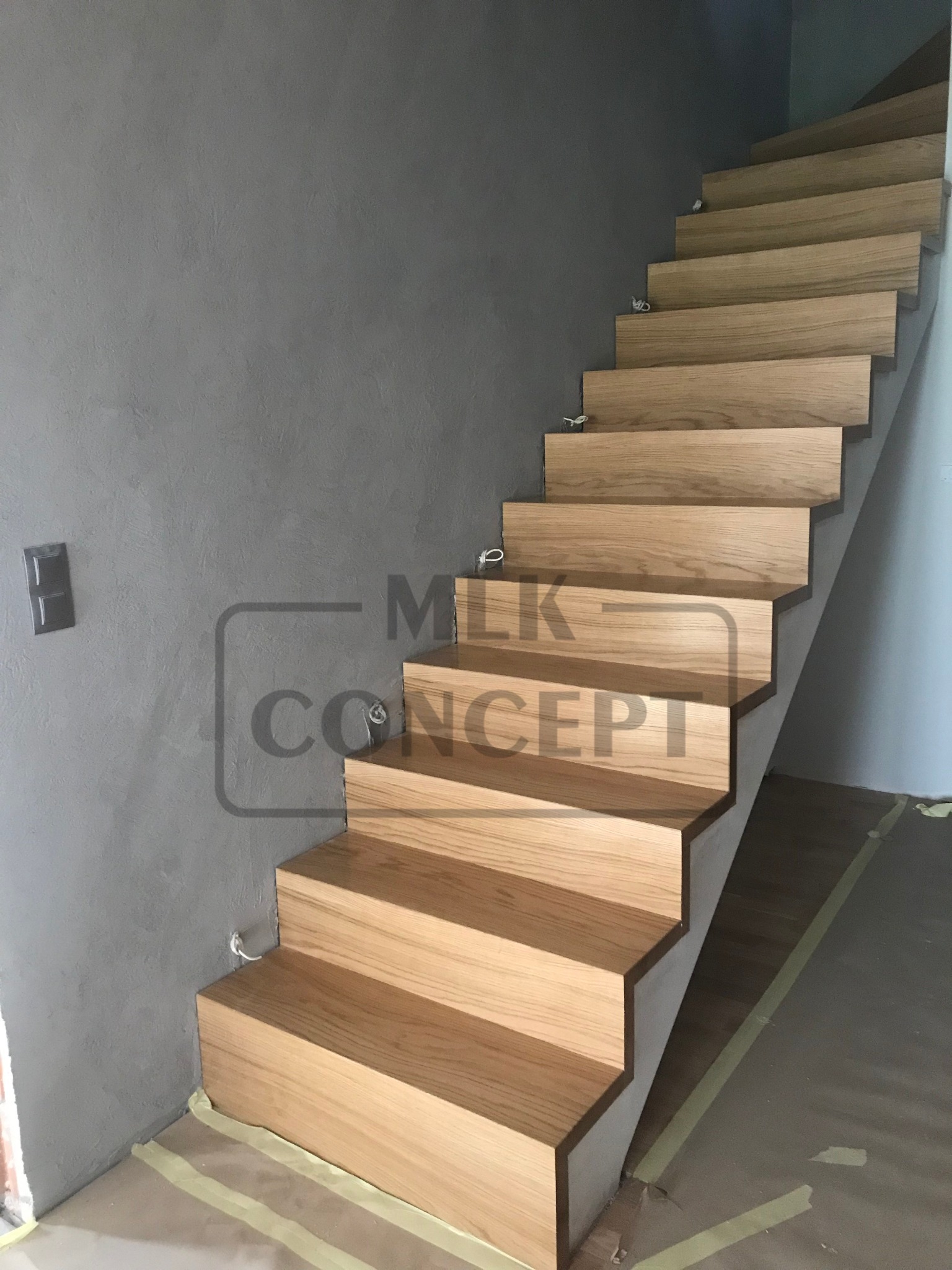 Proste, drewniane schody bez balustrady, z widocznymi przewodami elektrycznymi wystającymi ze ściany, zabezpieczone taśmą malarską na podłodze.