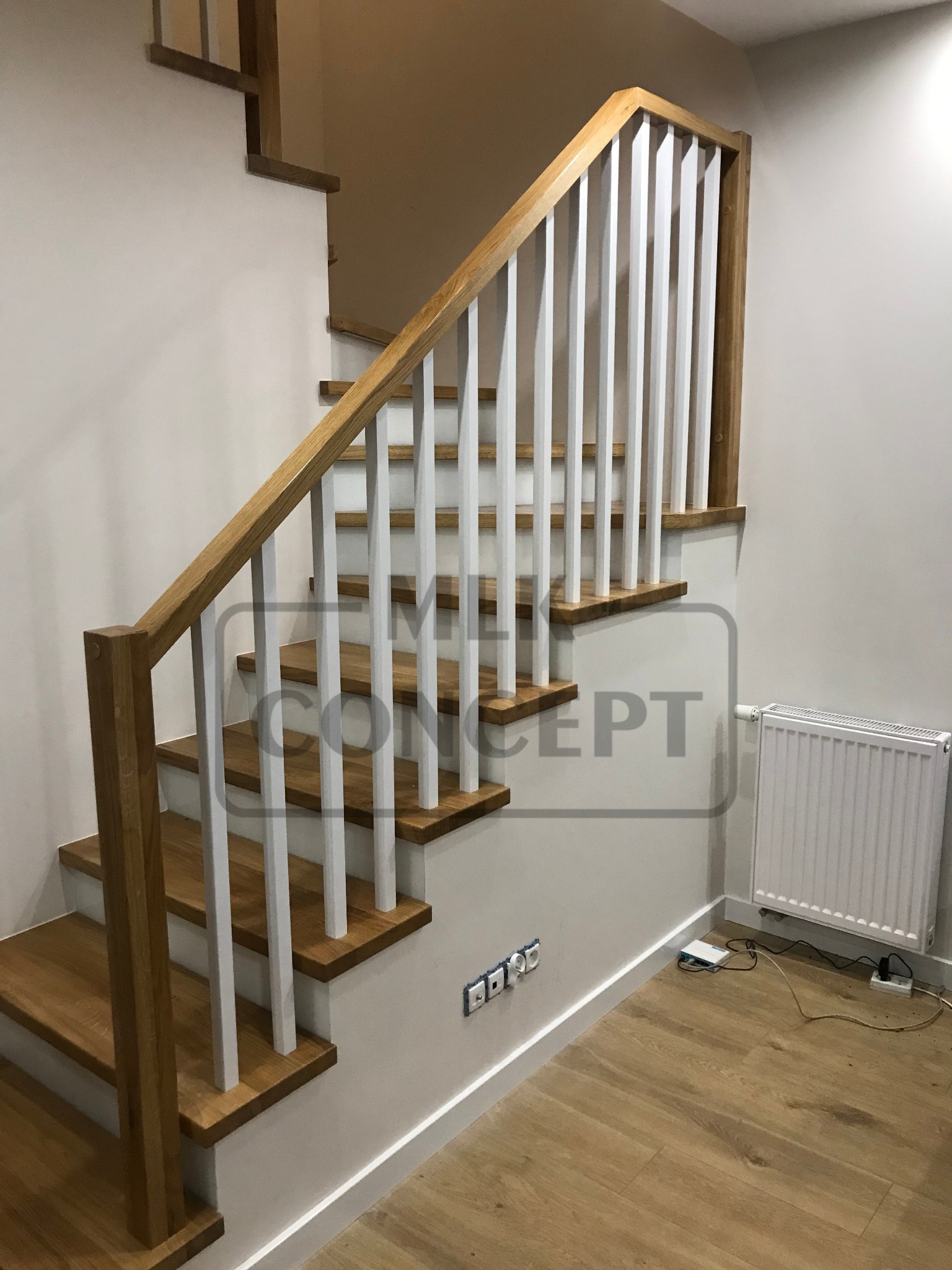 Jasne schody z drewnianymi stopniami i białymi tralkami, widok z dołu, z grzejnikiem i gniazdkami na ścianie w tle.