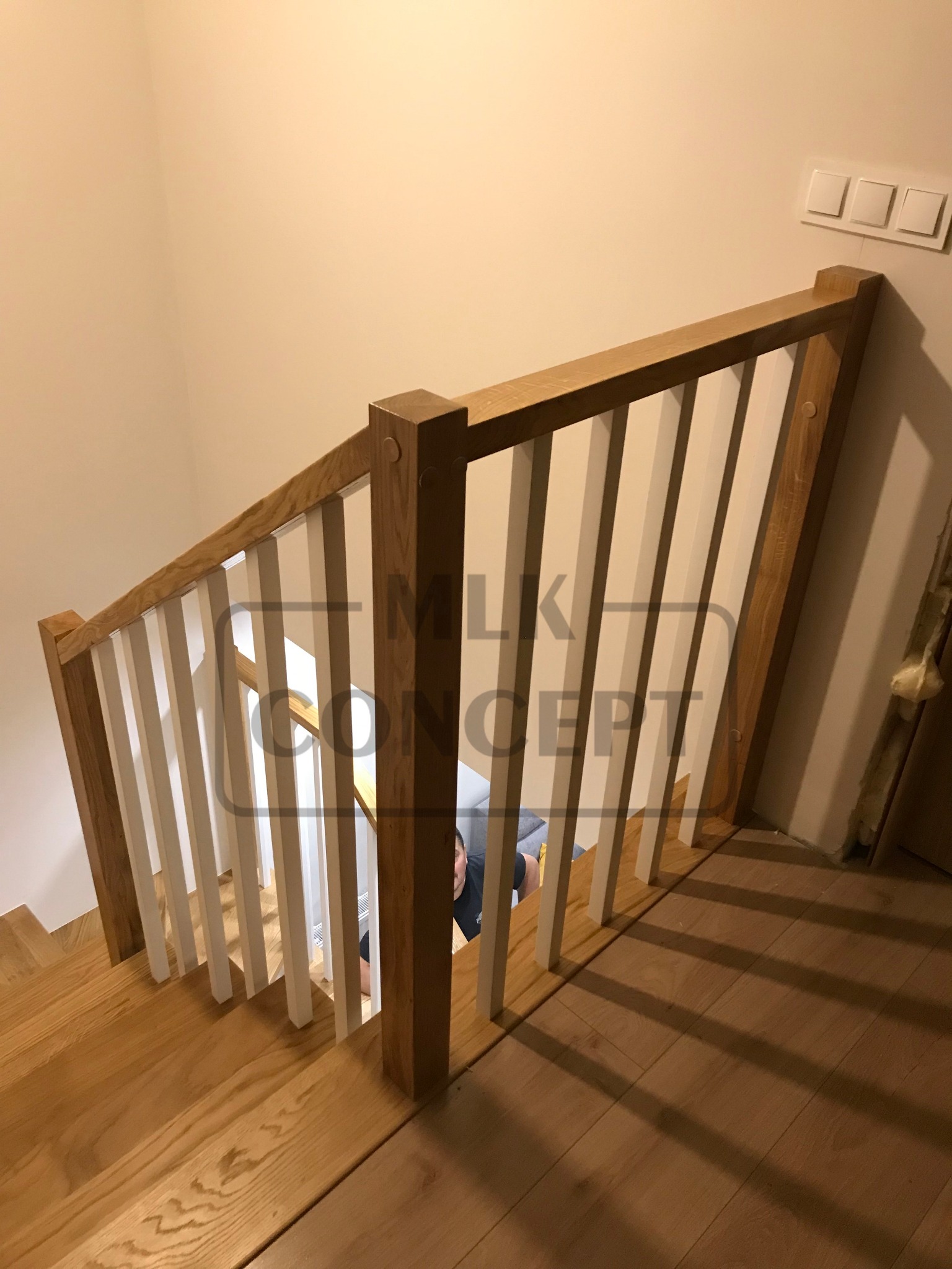 Widok z góry na drewniane schody z jasnymi drewnianymi poręczami i białymi szczeblami, na ścianie widoczne trzy białe włączniki światła.