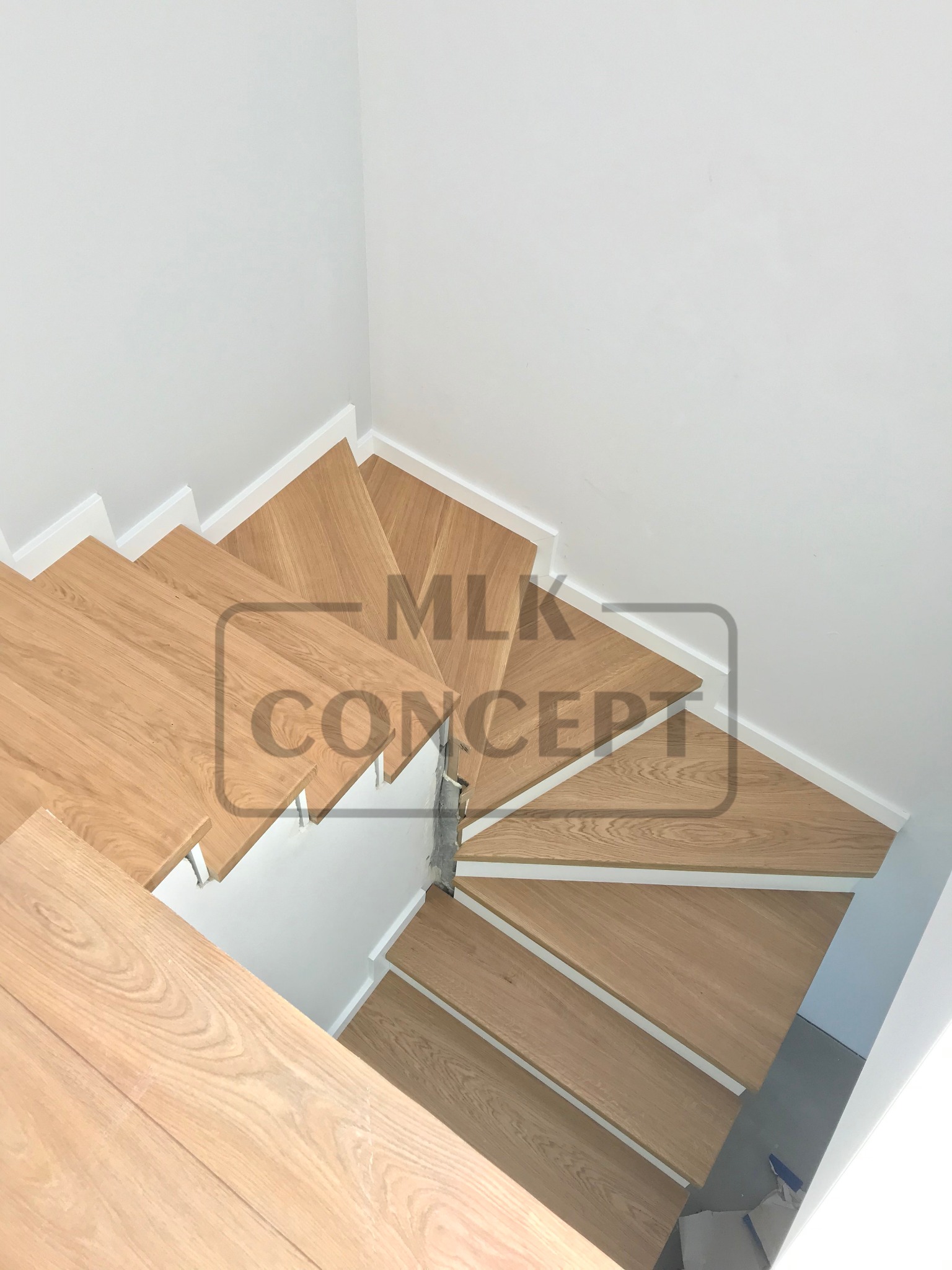 Widok z góry na skręcające jasne, drewniane schody z białymi podstopnicami, oświetlone naturalnym światłem, w minimalistycznym wnętrzu.