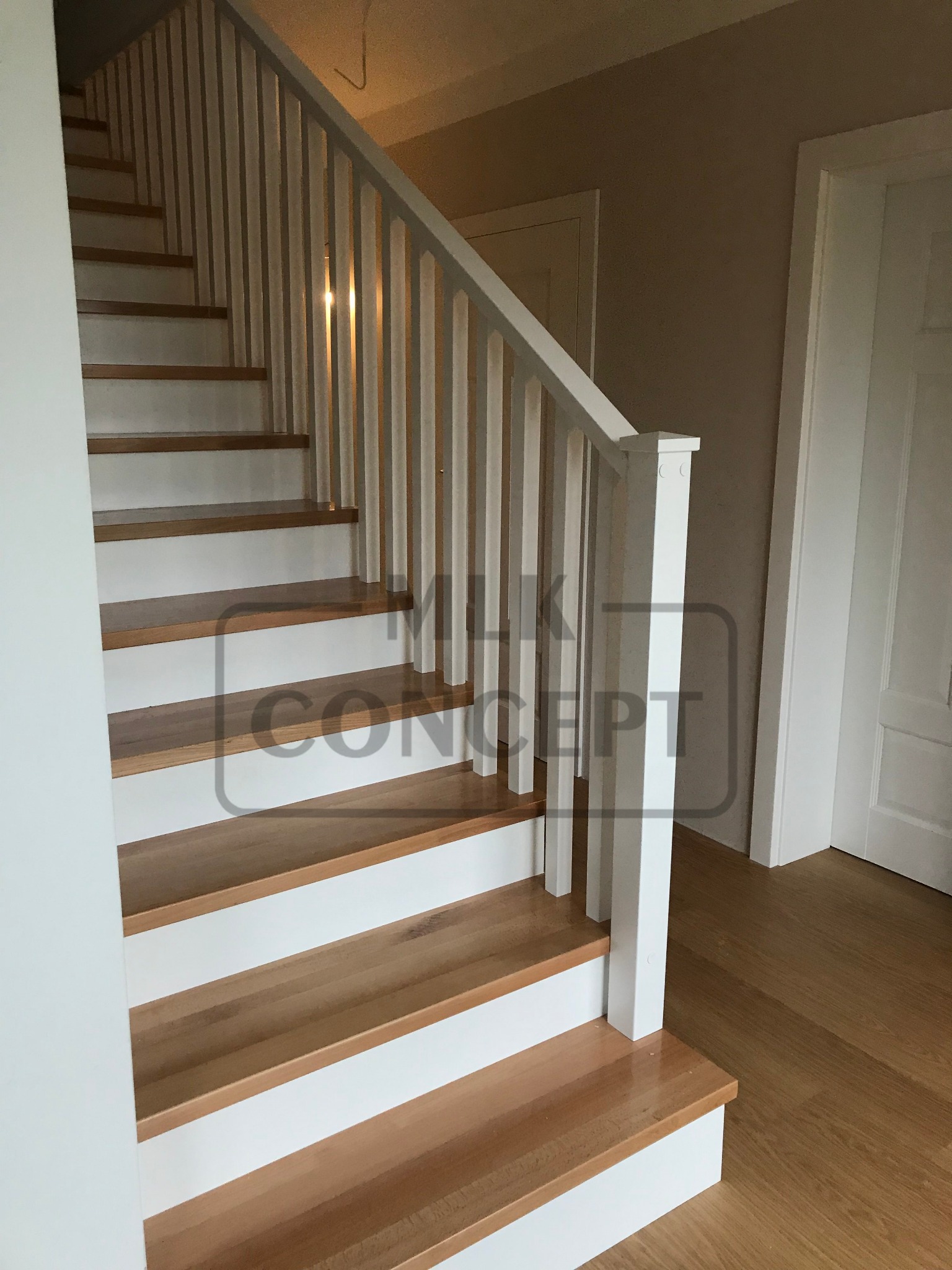 Jasne schody z drewnianymi stopniami i białymi podstopnicami oraz białą balustradą z pionowymi szczeblami w jasnym wnętrzu.