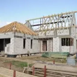 Budowa domu jednorodzinnego w stanie surowym otwartym: białe bloczki, drewniana konstrukcja dachu, widoczna betoniarka i składowane deski na placu budowy.