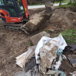 SajgonBUD usługi remontowo-budowlane - Mini koparka Kubota podnosi gruz i odpady budowlane na placu budowy. Widoczne ślady pracy w ziemi i nagromadzony gruz. Etap porządkowania terenu.