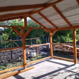 SajgonBUD usługi remontowo-budowlane - Drewniana konstrukcja zadaszenia tarasu w ogrodzie, widok z zewnątrz. Częściowo zadaszona konstrukcja z belek, w tle zieleń ogrodu i budynek.