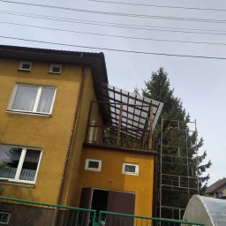 SajgonBUD usługi remontowo-budowlane - Żółty dom z dobudowanym balkonem z drewnianą konstrukcją zadaszenia. Widoczne rusztowanie i fragment szklarni. Ujęcie z dołu, pochmurne niebo.