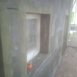 SajgonBud - Ocieplona ściana budynku z widocznym oknem zabezpieczonym folią. Fragment pomarańczowej taśmy leży na parapecie. Widok z boku, z bliska.