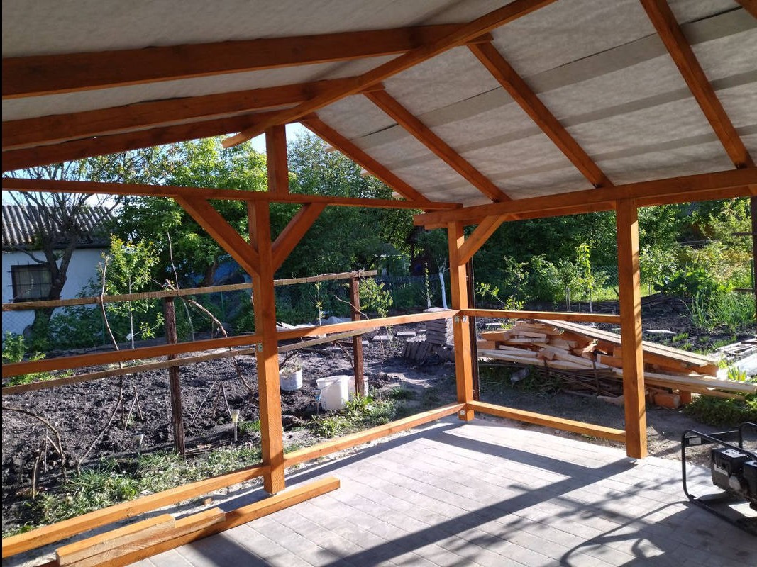 Drewniana konstrukcja zadaszenia tarasu w ogrodzie, widok z zewnątrz. Częściowo zadaszona konstrukcja z belek, w tle zieleń ogrodu i budynek.