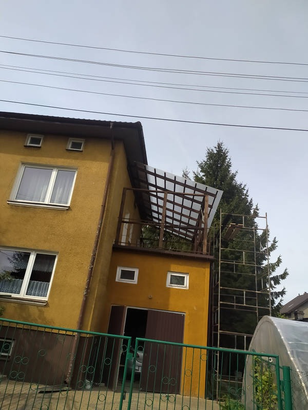 Żółty dom z dobudowanym balkonem z drewnianą konstrukcją zadaszenia. Widoczne rusztowanie i fragment szklarni. Ujęcie z dołu, pochmurne niebo.