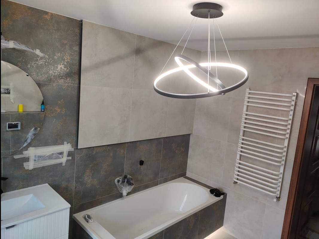 Nowoczesna łazienka w trakcie wykańczania: biała wanna, grzejnik drabinkowy, okrągłe lustro i designerska lampa. Widoczne zabezpieczenia na elementach.