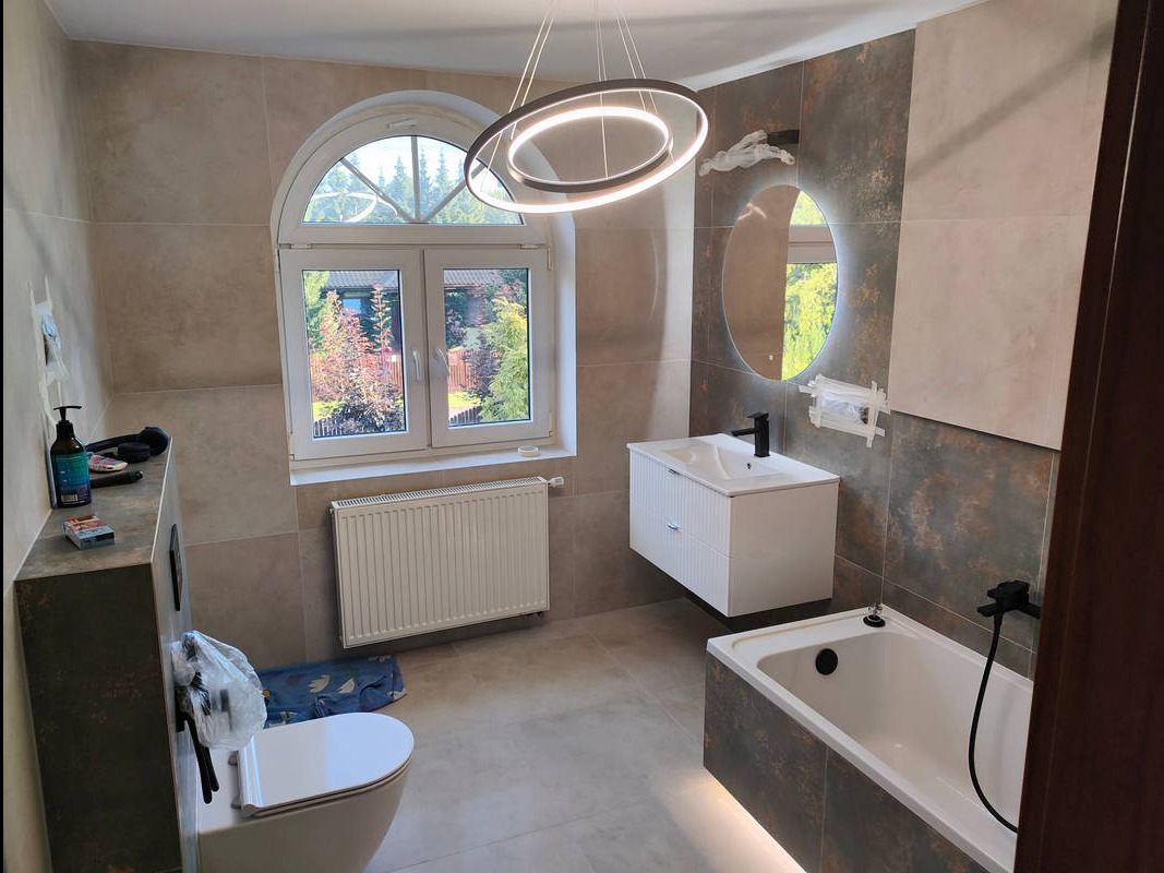 Nowoczesna łazienka z oknem łukowym: biała umywalka, okrągłe lustro z podświetleniem, grzejnik i wisząca lampa. Płytki w odcieniach beżu i brązu.