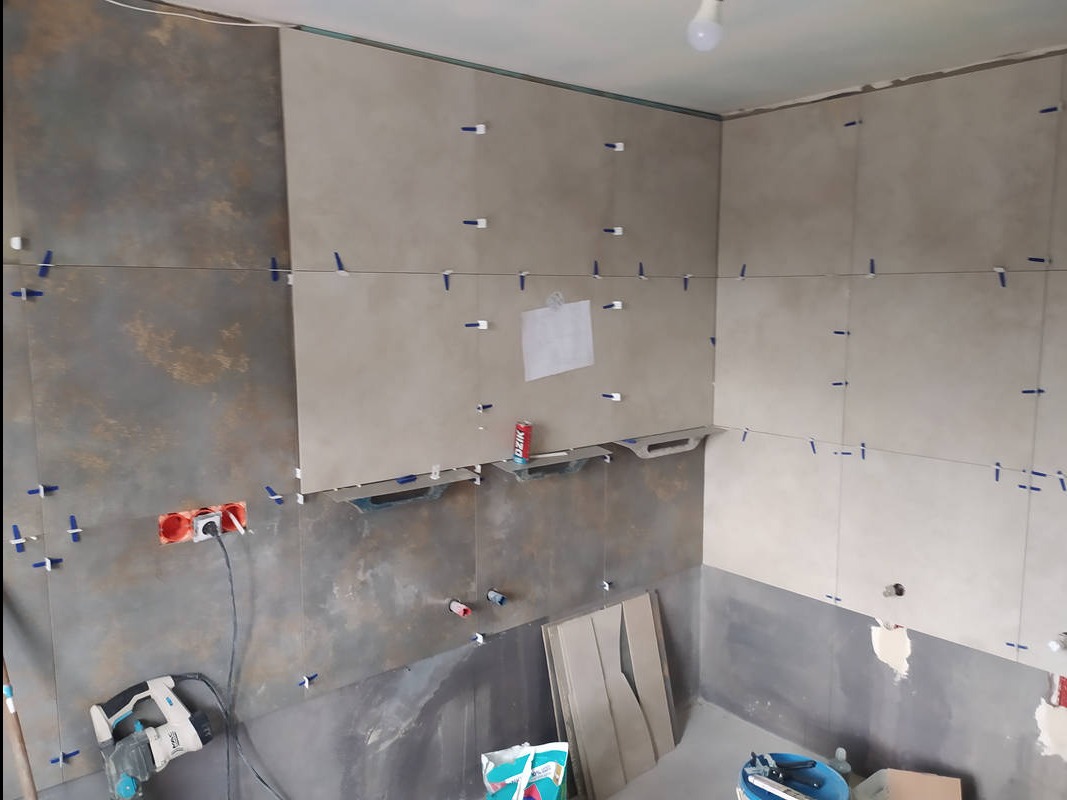 Wnętrze łazienki w trakcie układania płytek. Widoczne narzędzia, poziomica laserowa, krzyżyki dystansowe i częściowo ułożone płytki w odcieniach szarości i beżu.