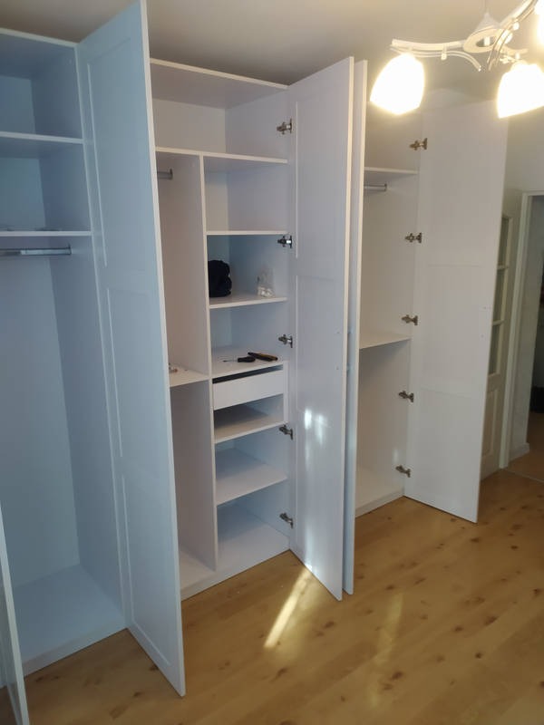 Biała, modułowa szafa wnękowa z otwartymi drzwiami, ukazująca półki i drążki na ubrania. Widoczna drewniana podłoga i fragment wnętrza mieszkania.